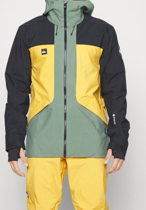 Herre regnjakke med farveblokke og hætte i sort, gul og grøn, kombineret med gule bukser, der har lynlås og tommelhul ved ærmekanterne.