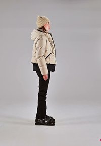 Vrouw die zijwaarts staat, draagt een beige donsjacket, bijpassende gebreide muts, zwarte broek en zwarte skischoenen op een grijze achtergrond.