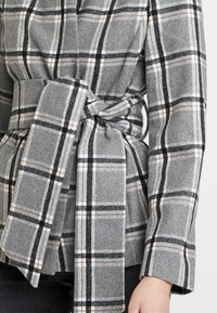 Manteau à carreaux gris avec une taille ceinturée, fabriqué en tissu texturé, avec des lignes noires et blanches et de subtils accents marron.