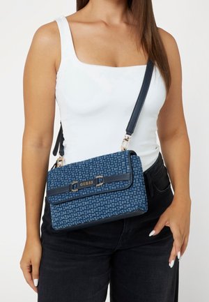 Kobieta w białym topie na ramiączkach i czarnych spodniach, z niebieską, teksturowaną torebką Guess na ramię.