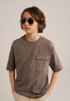 Nooruk laineliste pruunide juustega, kandes tumedaid päikeseprille, taupe'i värvi suure taskuga suurt T-särki ja beeže pükse, käed taskutes.