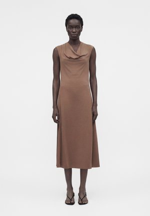 Γυναίκα στέκεται φορώντας αμάνικο καφέ μίντι φόρεμα με ριχτό ντεκολτέ και μαύρα ψηλοτάκουνα σανδάλια με λουράκια σε λευκό φόντο.