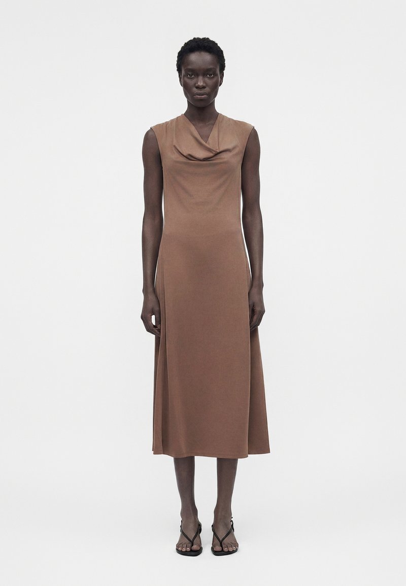 Γυναίκα στέκεται φορώντας αμάνικο καφέ μίντι φόρεμα με ριχτό ντεκολτέ και μαύρα ψηλοτάκουνα σανδάλια με λουράκια σε λευκό φόντο.