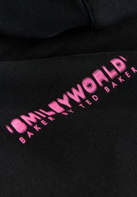 Musta kangas, jossa näkyvänä vaaleanpunainen kirjailuteksti "SMILEYWORLD BAKER BY TED BAKER." Sileä pinta, puhtaat linjat, moderni muotoilu.