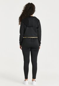 Zwarte sportkleding set bestaande uit een cropped hoodie met capuchon en gouden accent op de elastische tailleband, gestructureerde stof en nauwsluitende broek.