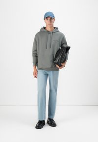 Grå hettegenser, lyseblå jeans, svarte sko. Grå ryggsekk holdes i venstre arm. Blå caps. Enkelt design, ingen synlige mønstre.