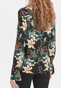 Kvinna med långt lockigt hår bär en svart blus med stort vitt och gult blommönster samt svarta byxor, visad bakifrån.