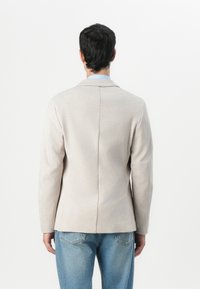 Hellbeige Blazer mit taillierter Passform, einreihiger Front, mit Revers und glatter Textur, getragen über einem blauen Hemd mit Kragen.