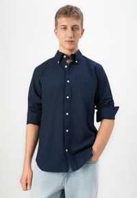 Joven con cabello corto rubio, vistiendo una camisa de botones azul marino con las mangas arremangadas y jeans de mezclilla de color azul claro, de pie frente a un fondo blanco.