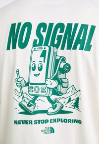 Hvid t-shirt med en grøn grafik af en tegneserie-smartphonekarakter med en rygsæk, på vandretur i et bjerglandskab. Tekst: "INGEN SIGNAL STOP IKKE UDFORSKNING."