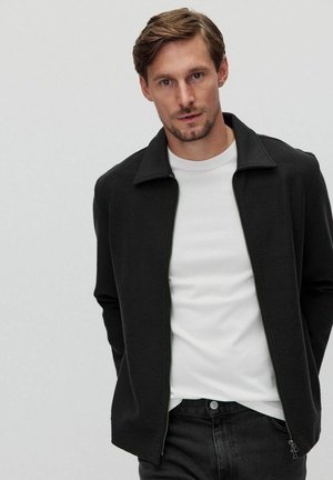 Black zip-up jacket with a textured fabric, a stand collar, and a slim fit, layered over a white crew-neck shirt and dark jeans.