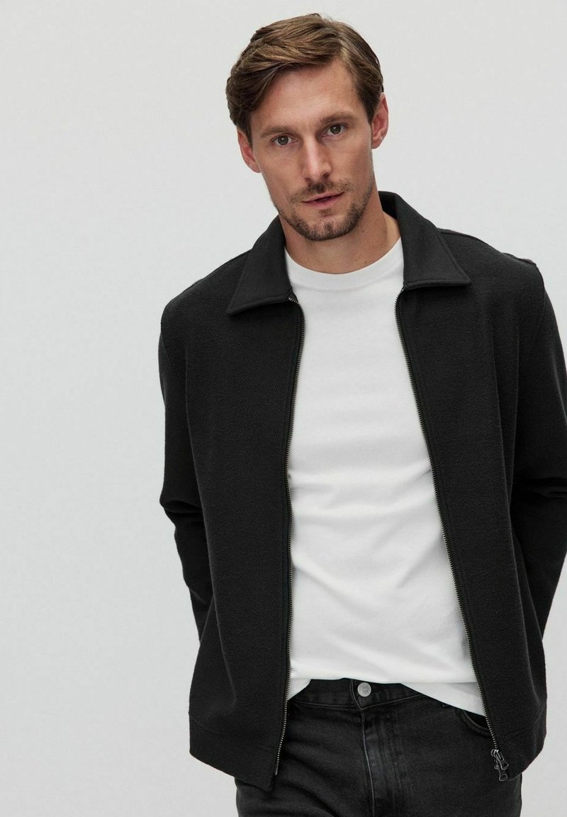 Sort lynlåsfleecejakke med struktureret stof, en opretstående krave og slim fit, lagdelt over en hvid crew-neck t-shirt og mørke jeans.