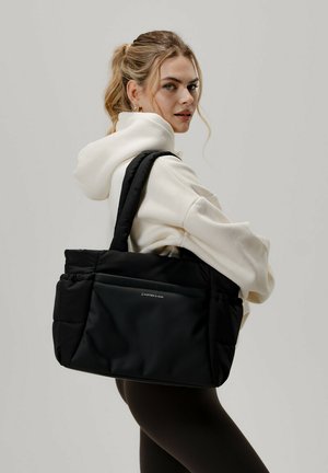 Bolso tote acolchado negro con asas dobles, que presenta una textura de tela suave y un bolsillo frontal con un logo. Diseño minimalista.