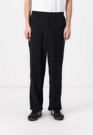 Zwarte ribbroek met een relaxte pasvorm, voorzien van een knoopsluiting en een gestructureerde stof. Gecombineerd met zwarte sneakers.