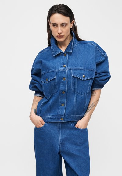 Άτομο με μακριά σκούρα μαλλιά που φοράει μπλε τζιν μπουφάν και ταιριαστό παντελόνι, με τα χέρια στις τσέπες, με ορατά τατουάζ στα πήχεις.