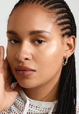 Pendientes de aro dorados con un diseño retorcido, combinados con un top de punto blanco texturizado. Enfocándose en las características faciales, incluyendo piel suave y cabello trenzado.