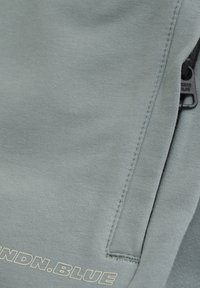 Ljusgrå sweatshirt med mjuk textur, synliga sömmar och en dragkedjeficka med en svart etikett som visar "LNDN.BLUE."