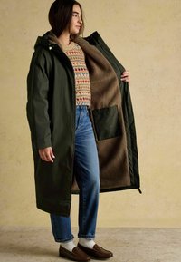 Lang olivengrøn frakke med en brun, fleeceforet inderside, udstyret med hætte og en forlommer. Båret over en mønstret sweater og blå jeans.