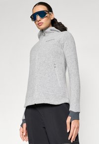 Veste en polaire grise avec capuche, fermeture éclair intégrale et poches latérales. Comprend des trous pour les pouces aux poignets et un tissu texturé pour la chaleur.