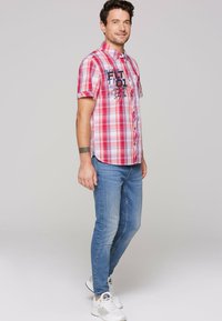 Kurze ärmellose karierte Hemd in Rot und Weiß mit Knopfschluss und Frontdruck. Kombiniert mit hellblauen Jeans und weißen Sneakern.