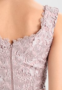 Roze jurk met bloemenkant overlay, golvende halslijn en ritssluiting aan de achterkant. Textuur borduursel voegt detail toe.