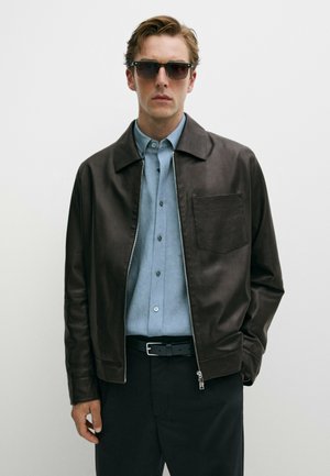 Homme portant une veste en cuir marron foncé, une chemise bleu clair boutonnée, un pantalon noir, une ceinture noire et des lunettes de soleil foncées sur un fond uni.
