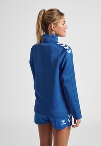 Blauwe sportjasje met hoge kraag en lange mouwen, voorzien van witte chevronpatronen op de schouders. Gecombineerd met blauwe shorts van glad materiaal.