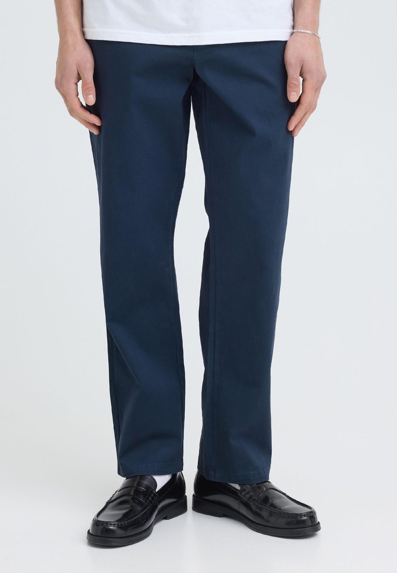 Personne portant un pantalon bleu marine droit, des chaussettes blanches, des mocassins en cuir noirs et une chemise blanche, debout devant un fond uni.