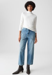 Weißes, enganliegendes Langarmoberteil, hellblaue hochgeschnittene, weit geschnittene Jeans und schwarze, spitze Stiefeletten. Glatte Textur und klare Linien.