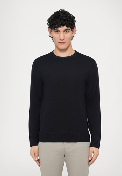 Black knitted crew neck sweater with long sleeves, ribbed cuffs, and hem, worn over light-colored pants against a plain background.