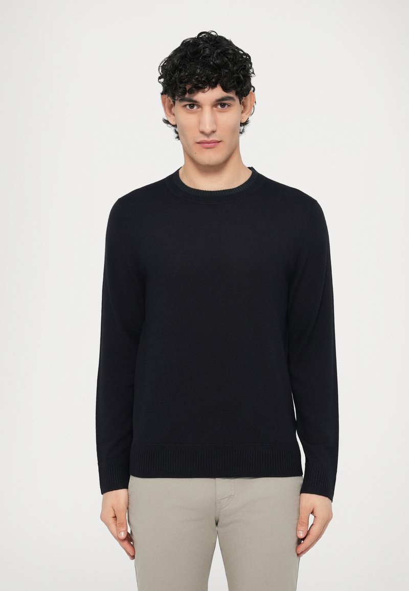 Czarny dziergany sweter z okrągłym dekoltem, długimi rękawami, ściągaczami na mankietach i u dołu, noszony na jasnych spodniach na jednolitym tle.