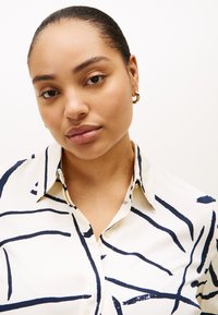 Πουκάμισο με κουμπιά από εκρού ύφασμα με σκούρες μπλε αφηρημένες γραμμές, γιακά και εμπρόσθιο πατιλέτα. Χρυσά σκουλαρίκια κρίκοι προσθέτουν μεταλλική πινελιά.