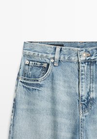 Lichtblauwe spijkerbroek taillegedeelte met voorste knoop, riemlussen, muntenzakje en zichtbare stiksels op een witte achtergrond.