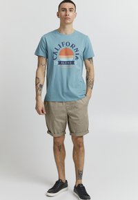Lyseblå bomulds T-shirt med "CALIFORNIA" grafik, kombineret med opsmøgede khaki shorts og sorte sneakers. Tatoveringer synlige på arme og ben.