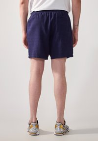 Shorts bleu marine en tissu texturé, avec une taille élastique et une poche arrière, associés à des chaussures de sport grises et jaunes.