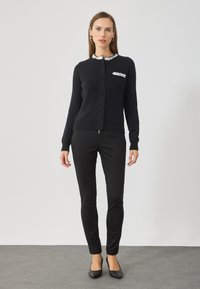 Czarny sweter z dzianiny z okrągłym kołnierzykiem, ozdobiony białymi cekinami, z zapięciem na guziki, noszony na czarnych dopasowanych spodniach i czarnych balerinach.