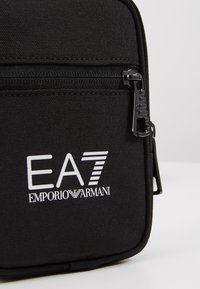 Czarna torba na zamek błyskawiczny wykonana z teksturowanej tkaniny, z białym logo "EA7 EMPORIO ARMANI" oraz czarnym zamkiem z polem do chwytania.