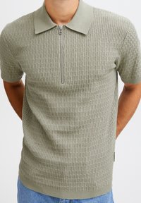 Khaki kortermet polo-skjorte med teksturert mønster, glidelås, og klassisk krage. Ribbet kanting og sidesplitt detaljer.