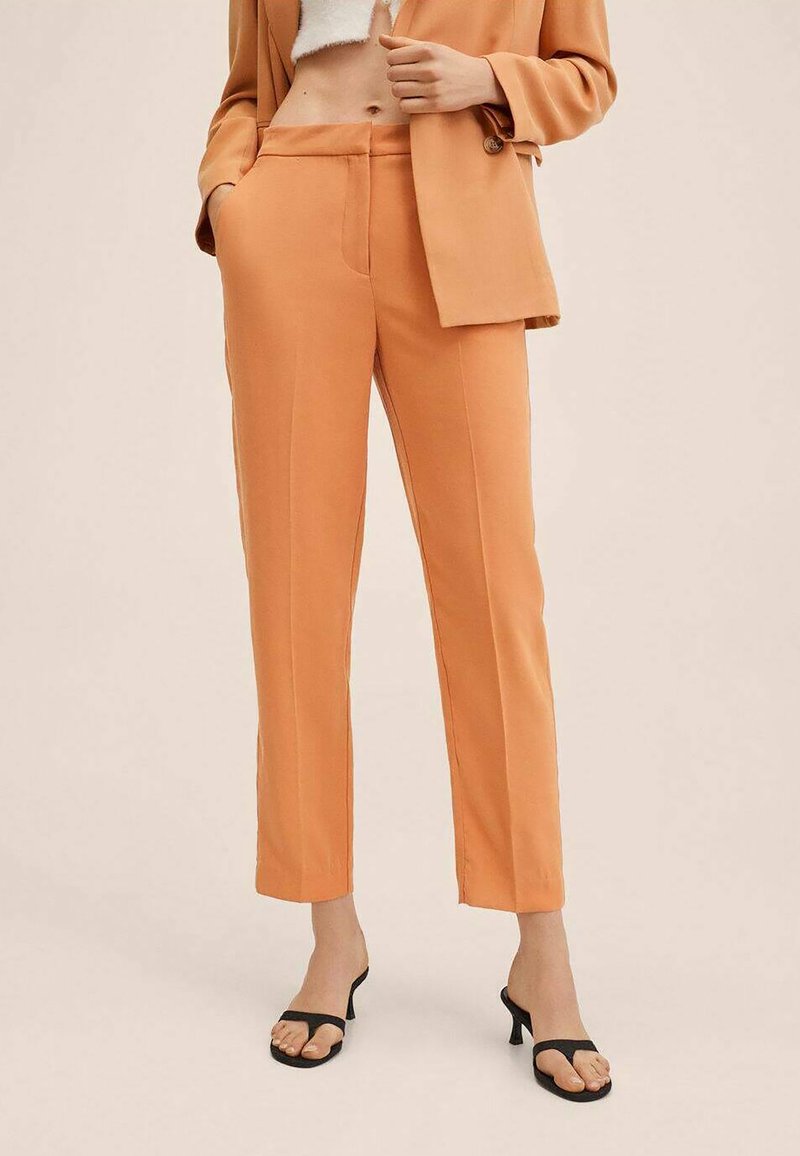 Femme portant un pantalon orange taille haute et ajusté avec poches latérales, associé à un blazer assorti et des sandales à talons ouvertes noires.