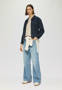 Giacca cropped blu navy, top beige a righe, jeans a gamba larga blu chiaro, maglione crema legato in vita, sneakers grigie con suola beige.