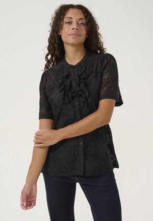 Zwarte kanten shirt met korte mouwen en een geraffineerde voorkant, met bloemmotieven en knoopsluitingen. Gecombineerd met donkere jeans.