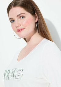 Camiseta blanca de cuello en V con letras de strass verdes que dicen "PRIMAVERA". Modelo con cabello largo y castaño, usando grandes aretes de plata.