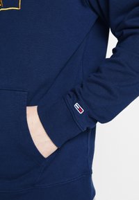 Tmavě modrá mikina s klokánkou, žebrovanými manžetami a vyšitým logem na rukávu. Vyrobena z měkké, strukturované látky.