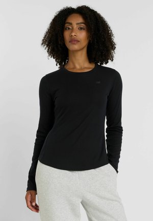 Haut noir à manches longues en tissu lisse et extensible, avec un col rond et un petit logo sur la poitrine ; associé à un pantalon de survêtement gris.