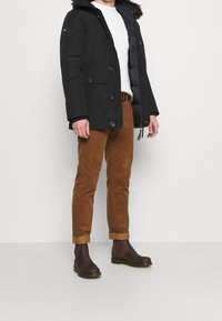Černá zateplená parka s kapucí lemovanou umělou kožešinou, se zapínáním na knoflíky a zip, nošena přes bílý svetr a hnědé manšestrové kalhoty.