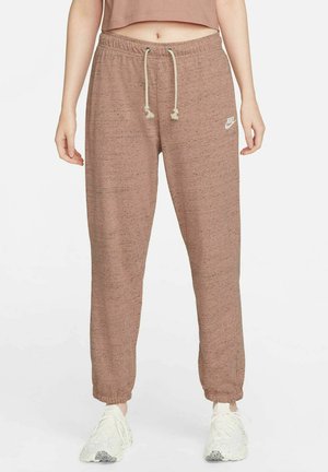 Frau trägt hellbraune Nike-Jogginghosen mit Kordelzug und weiße Turnschuhe, steht vor einem einfachen weißen Hintergrund.