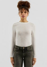 Weißes, langärmliges, geripptes Shirt mit kleinen silbernen Verzierungen und rundem Ausschnitt, kombiniert mit hoch taillierten dunklen Jeans.