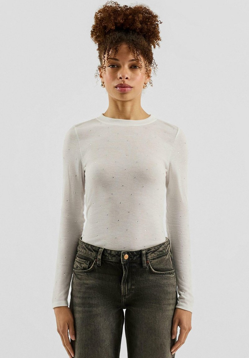 Weißes, langärmliges, geripptes Shirt mit kleinen silbernen Verzierungen und rundem Ausschnitt, kombiniert mit hoch taillierten dunklen Jeans.