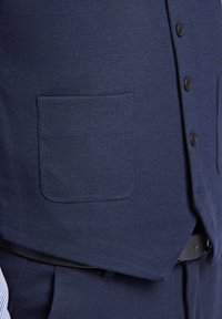 Dunkelblaues Weste mit einer Vorderseite Tasche, Knopfverschluss und einem strukturierten Stoff; getragen über einem hellen Hemd.