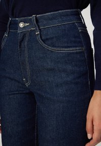 Jeans in denim blu scuro con vita alta, caratterizzati da cuciture visibili, tasche anteriori e chiusura con bottone metallico. Tessuto liscio.